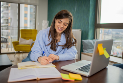 a young woman is writing in her notepad with an open laptop and book in front of her